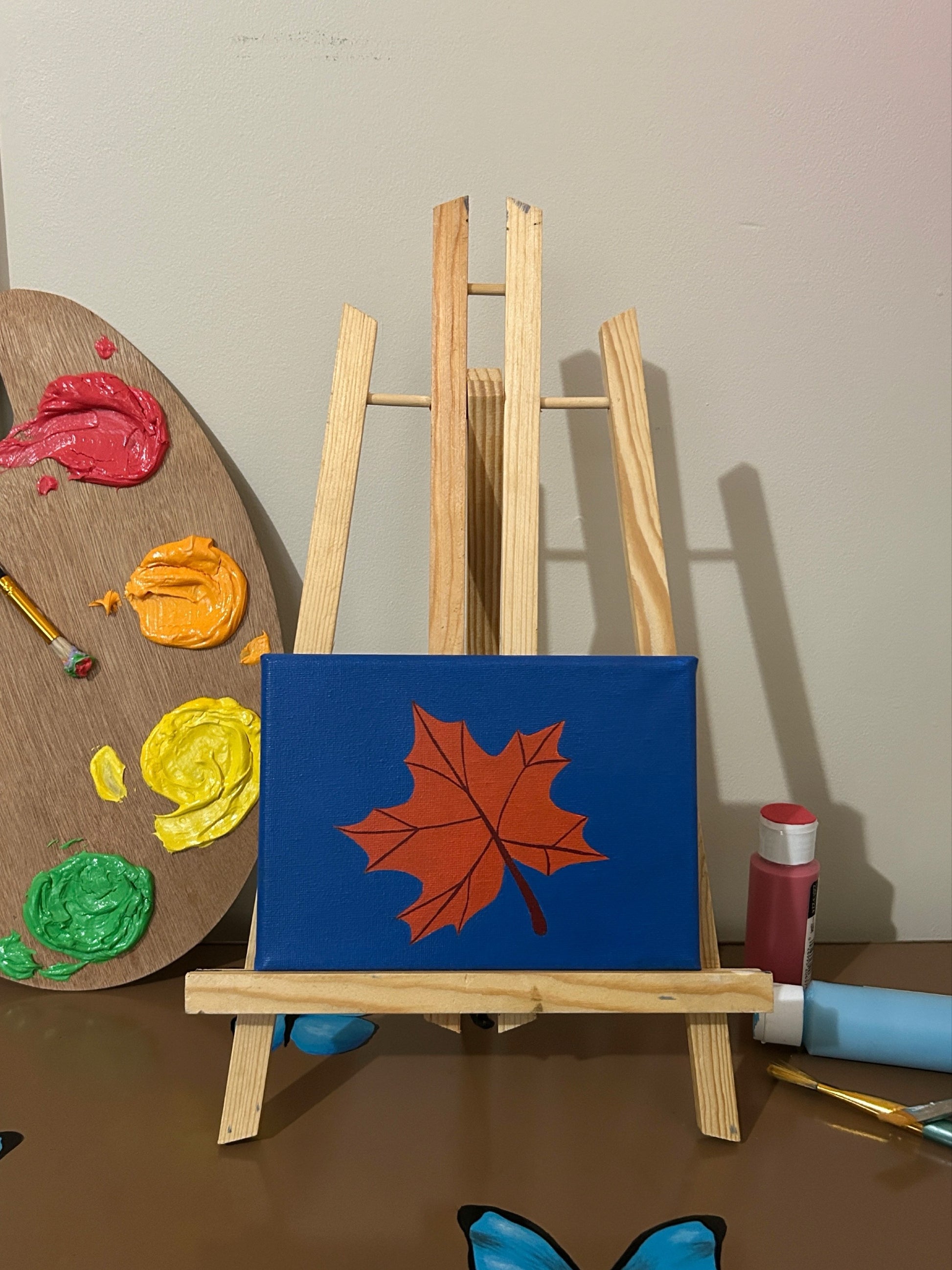 Amber Peeks Art: Small easel with a blue canvas featuring a orange leaf, surrounded by art supplies on a brown surface.
