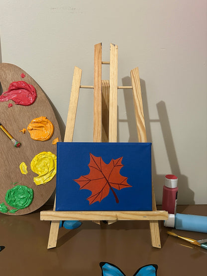 Amber Peeks Art: Small easel with a blue canvas featuring a orange leaf, surrounded by art supplies on a brown surface.