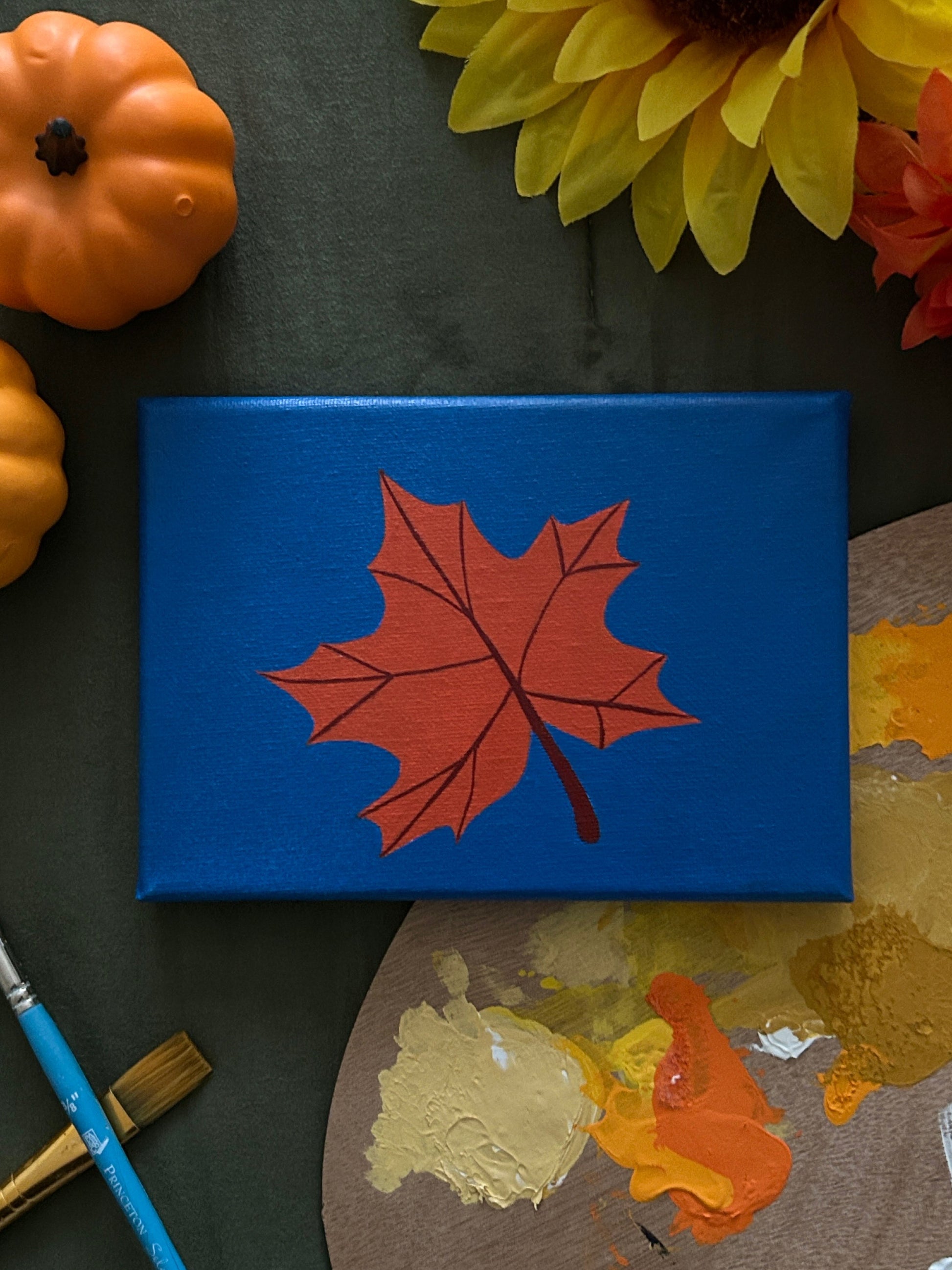 Amber Peeks Art: Blue canvas with a orange maple leaf on a dark surface with paint and pumpkins.