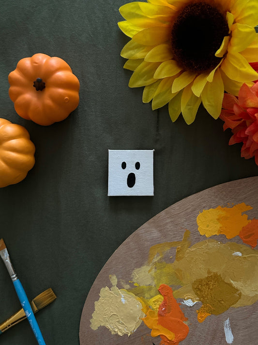 Amber Peeks Art: Small white ghost magnet on a dark surface with pumpkins, sunflowers, and a paint palette.