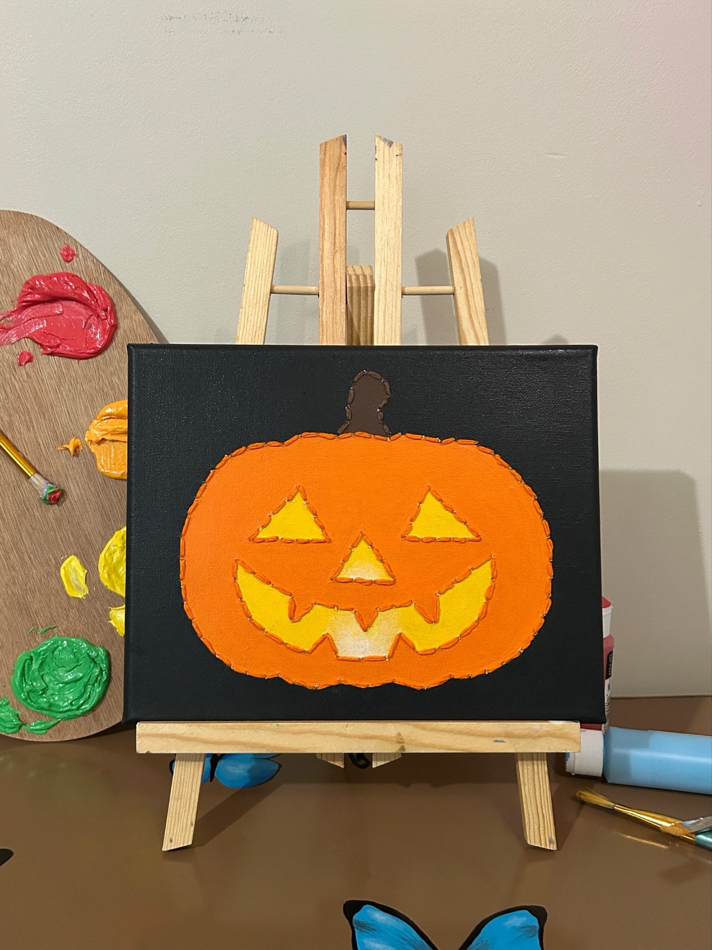Amber Peeks Art: painting of a jack-o'-lantern on a small easel with art supplies in the background.