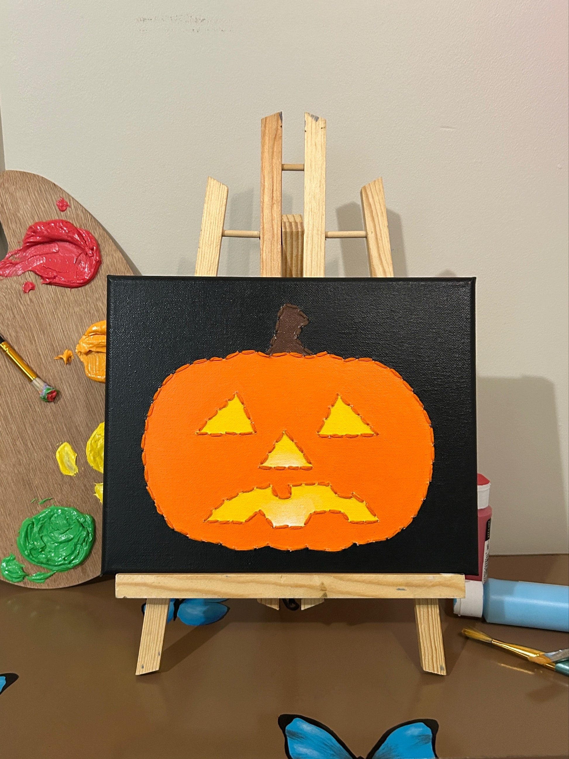 Amber Peeks Art: Children's painting of a jack-o'-lantern on a small easel with art supplies in the background.