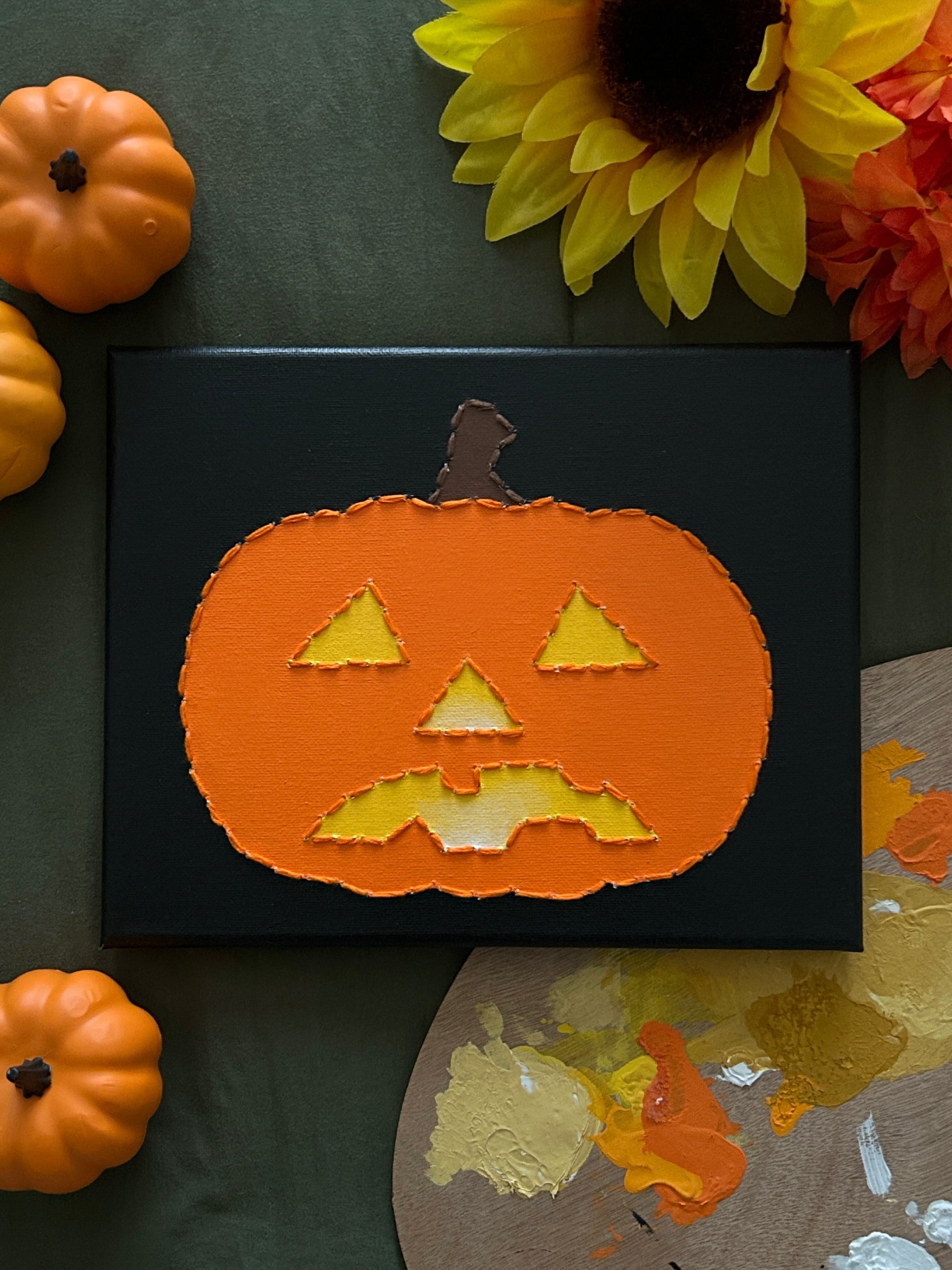 Amber Peeks Art: Decorative painting of a pumpkin with a face on a black background, surrounded by small pumpkins and sunflowers.
