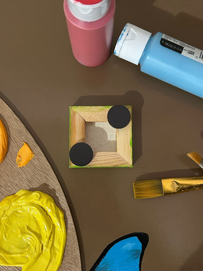 Amber Peeks Art: Artistic depiction of a kiwi slice on a magnet with painting supplies around it on a brown surface