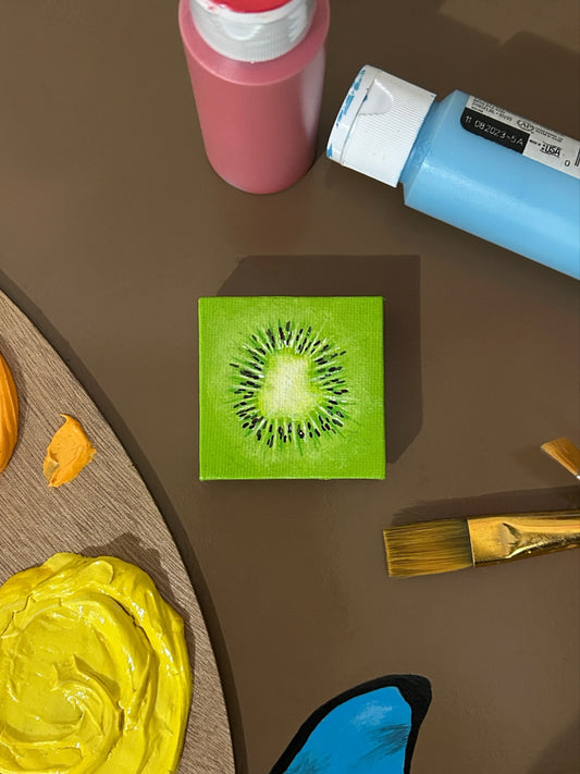 Amber Peeks Art: Artistic depiction of a kiwi slice on a magnet with painting supplies around it on a brown surface
