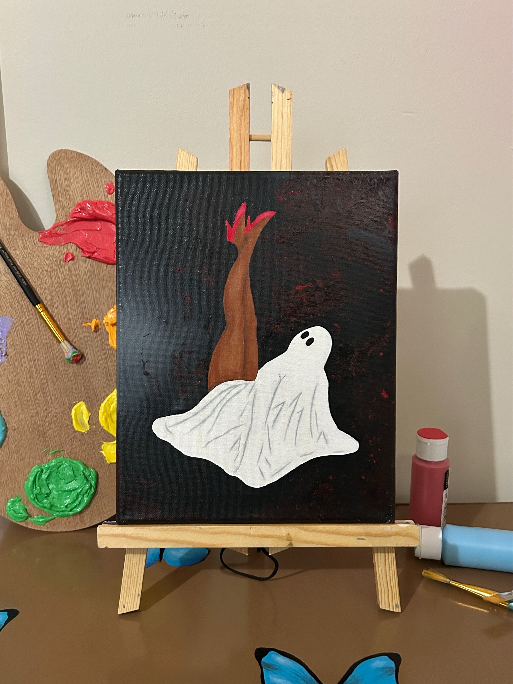 Amber Peeks Art: Painting of a ghost on a black background with a wooden easel, surrounded by art supplies.