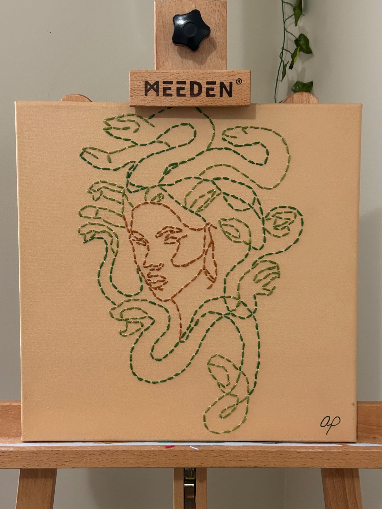 Amber Peeks Art: Embroidery of medusa with green thread on a beige canvas, displayed on a wooden easel.