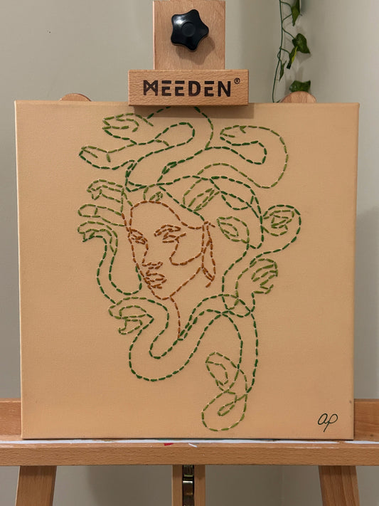 Amber Peeks Art: Embroidery of medusa with green thread on a beige canvas, displayed on a wooden easel.