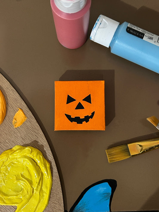 Amber Peeks Art: Small orange magnet with a pumpkin face design on a brown surface with art supplies.