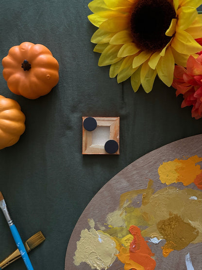 Amber Peeks Art: Small orange pumpkin magnet on a dark surface with pumpkins, sunflowers, and a paint palette.