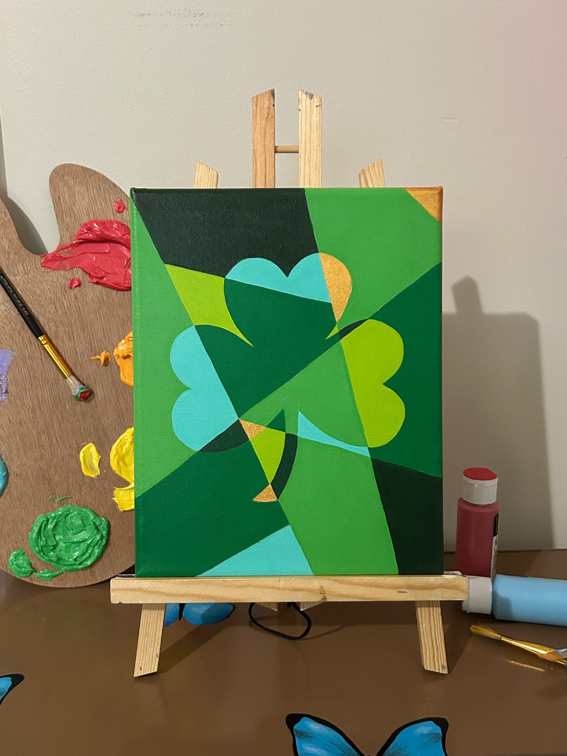 Amber Peeks Art: Green abstract painting of a shamrock on a small easel with art supplies in the background.