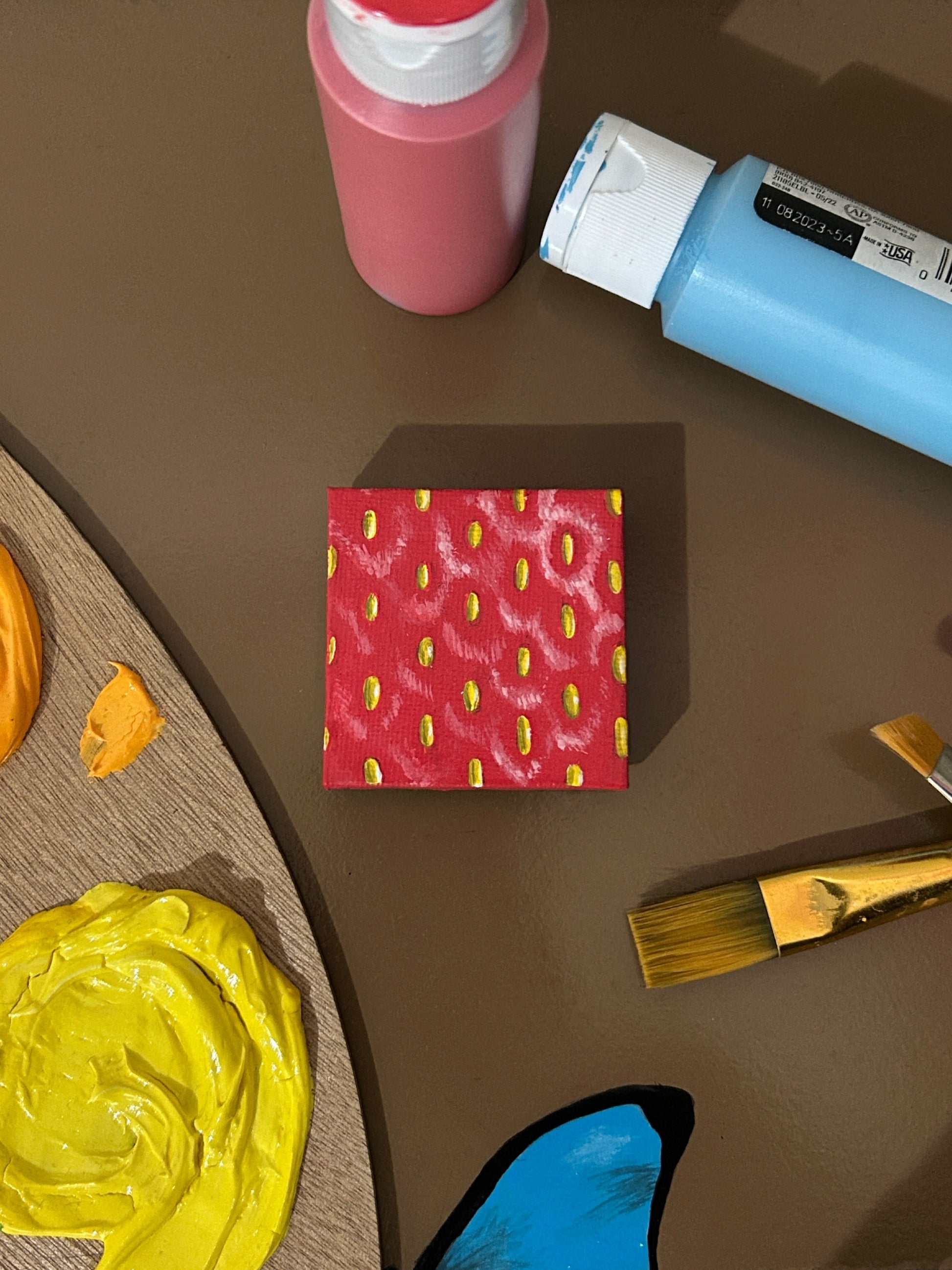 Amber Peeks Art: Small red magnet painted like a strawberry on a brown surface with art supplies.