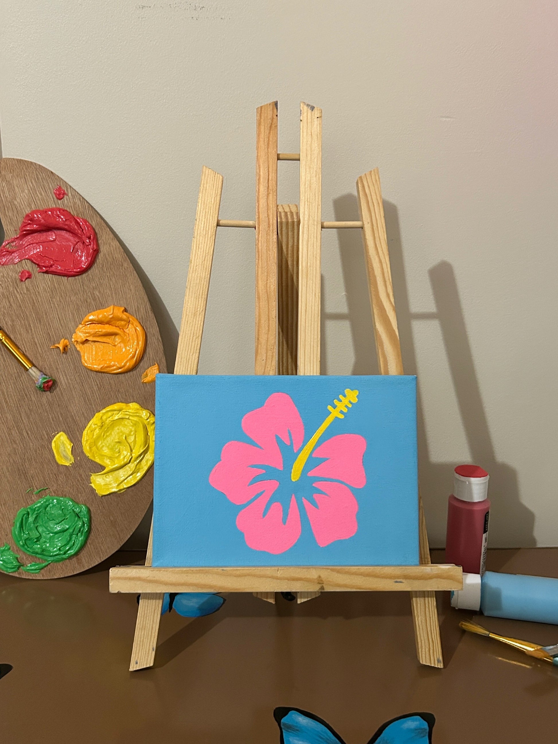 Amber Peeks Art: Canvas with a pink flower and blue background on a small easel next to a color palette and paintbrushes.